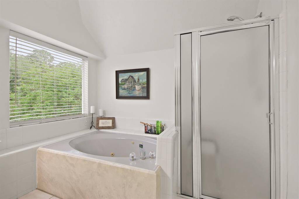 This primary bathroom features a large soaking tub with jets, a separate glass-enclosed shower, and a large window providing natural light and a view of greenery.