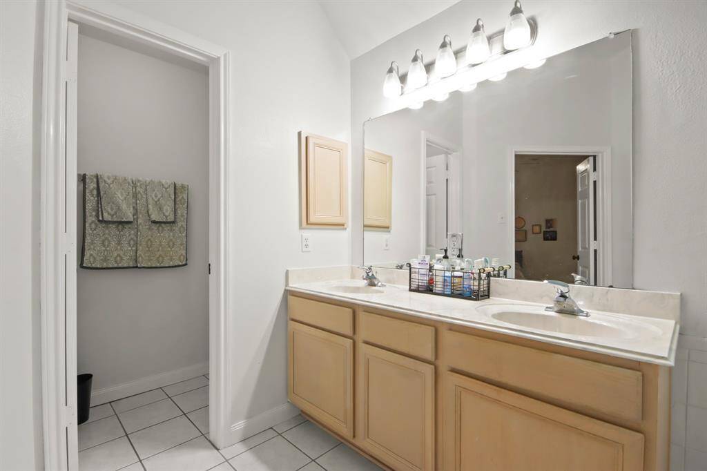 This photo shows a bright, spacious bathroom featuring a double sink vanity with ample storage, a large mirror, and modern lighting. It has a tiled flooring, offering a clean and inviting atmosphere.