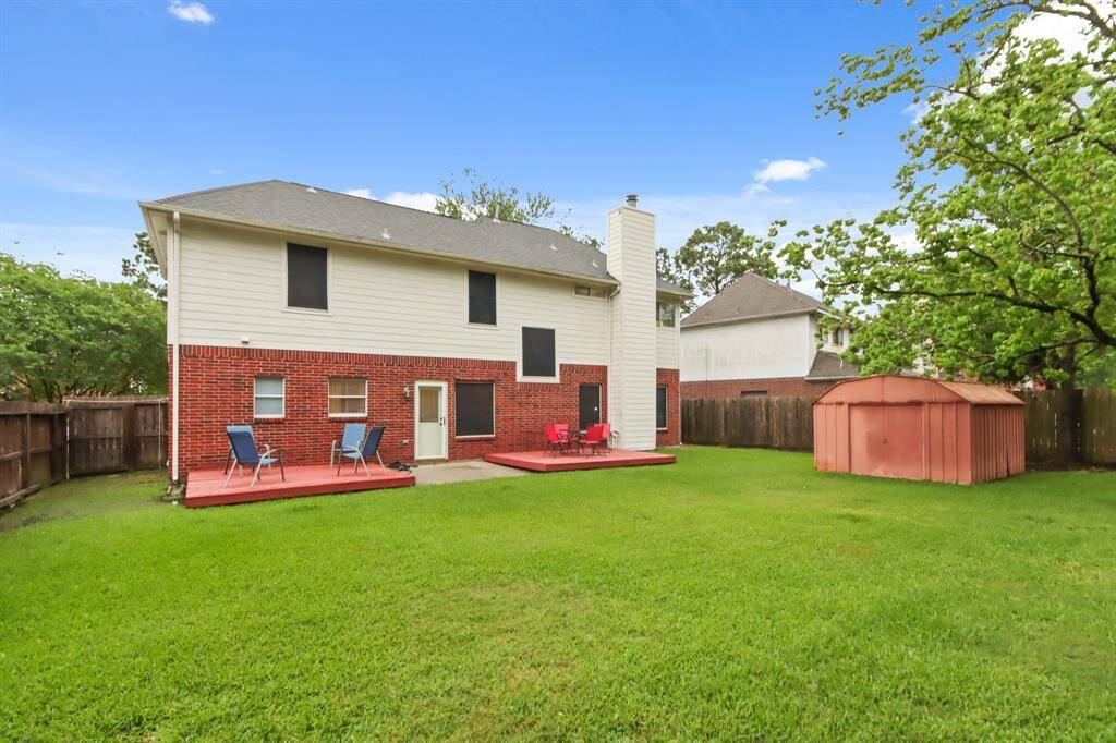 Spacious backyard with a red brick and siding two-story home, featuring a large lawn, a wooden deck/patio areas and a storage shed, all enclosed by a wooden fence.