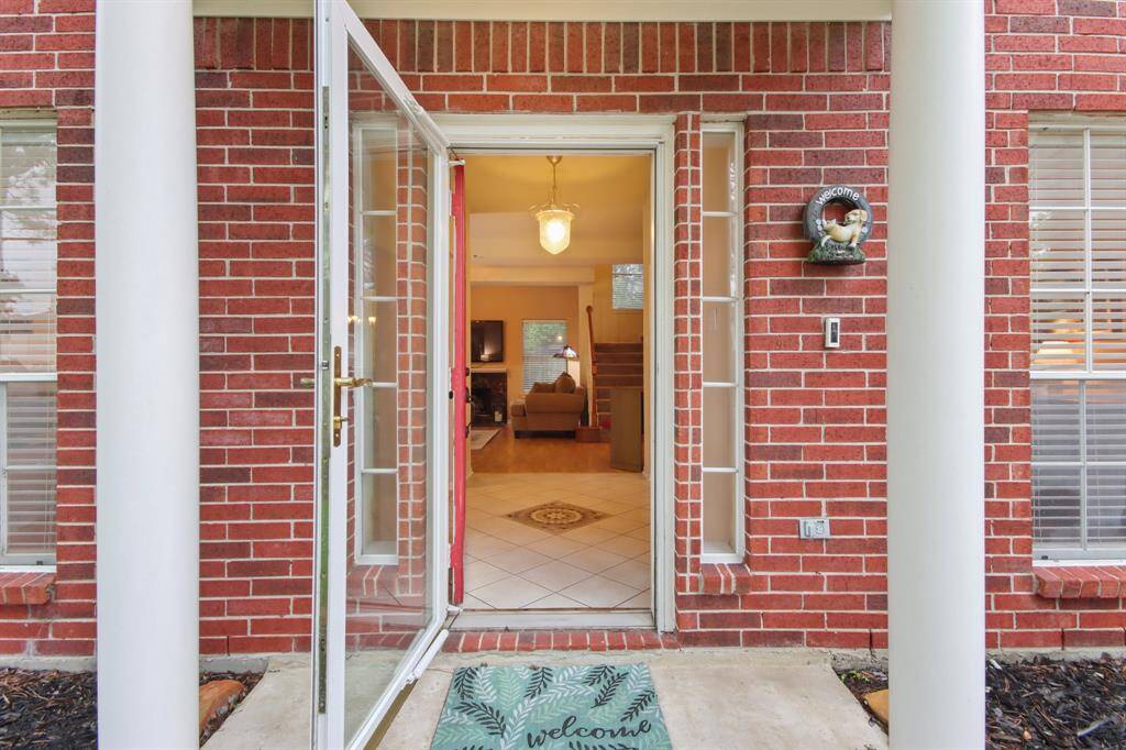 This welcoming covered entrance features a classic red brick exterior with white columns and a glass storm door. Inside, the view reveals a cozy living space with tiled flooring creating an inviting atmosphere.