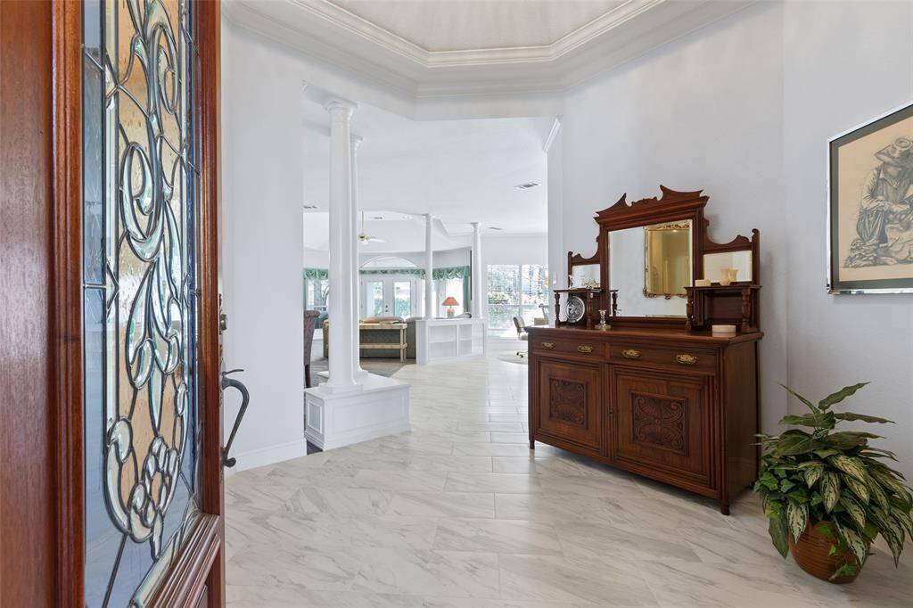 An elegant formal entry welcomes guests to this stunning lakeside property. Neutral tile floors sit beneath a sparkling chandelier, while tall windows in the background frame breathtaking views of the serene lake and lush greenery.