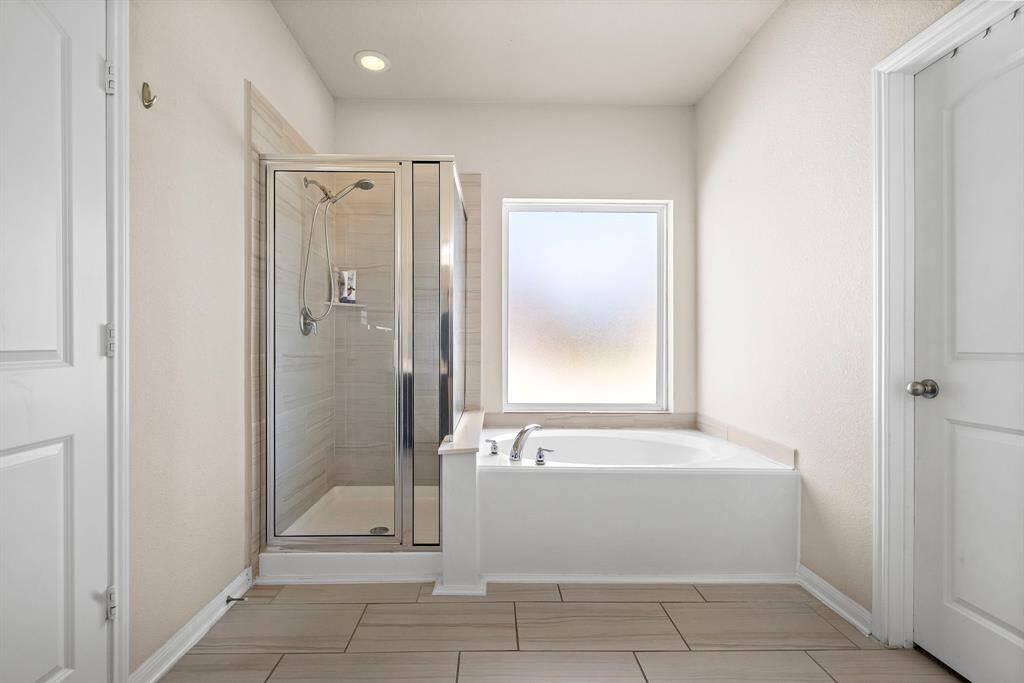 This primary bathroom features a modern design with a spacious glass-enclosed shower and a separate bathtub. It has neutral tones, tiled flooring, and a large frosted window for natural light while maintaining privacy.