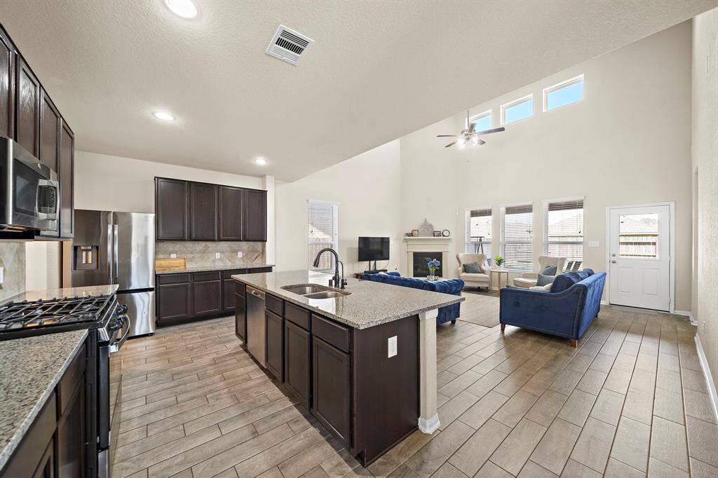 This open-concept living area features a modern kitchen with dark cabinetry, stainless steel appliances, and a large island. It flows into a spacious living room with high ceilings, abundant natural light, and a cozy seating area, perfect for family gatherings.