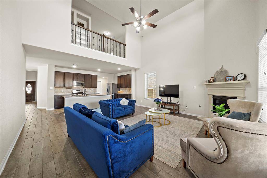 This spacious living area features a high ceiling and an open floor plan. It includes a cozy fireplace, modern kitchen with dark wood cabinets, and a loft-style second-floor overlooking the living space. Natural light fills the room, highlighting the stylish furnishings and wood-like flooring.