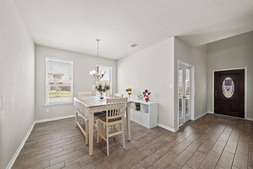 This photo showcases a bright dining area with wood-look tile flooring. It features a table set for six under a tasteful chandelier. Large windows provide ample natural light, and there's a stylish entryway with double glass doors nearby.