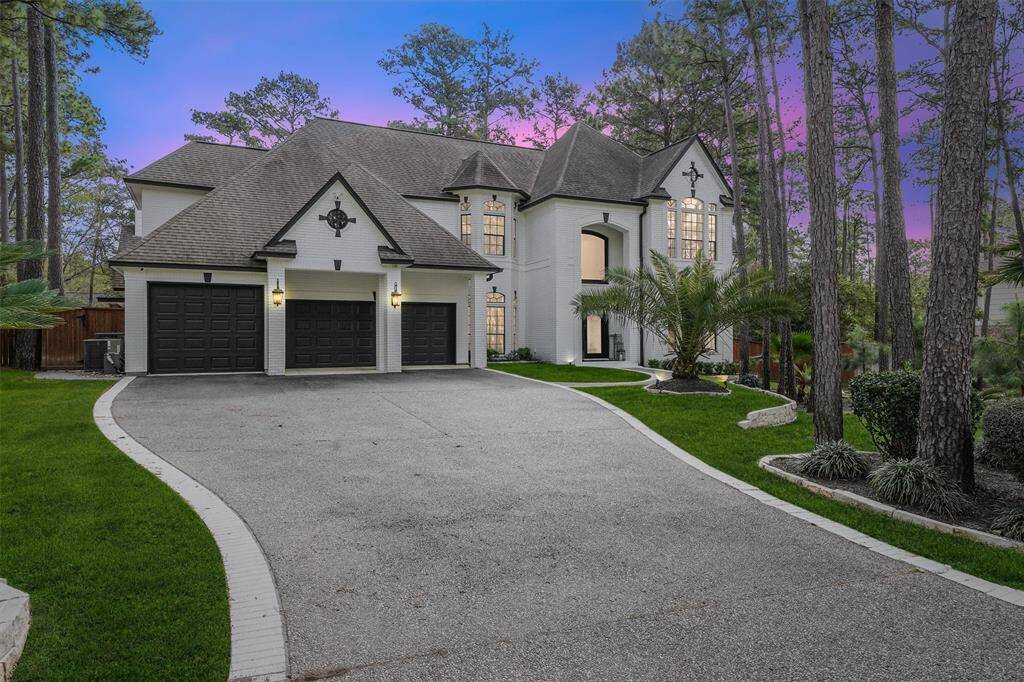 This elegant two-story home features a spacious driveway leading to a three-car garage, surrounded by lush greenery and tall trees. The exterior is highlighted by large windows and a beautifully landscaped front yard, perfect for a serene, wooded setting.