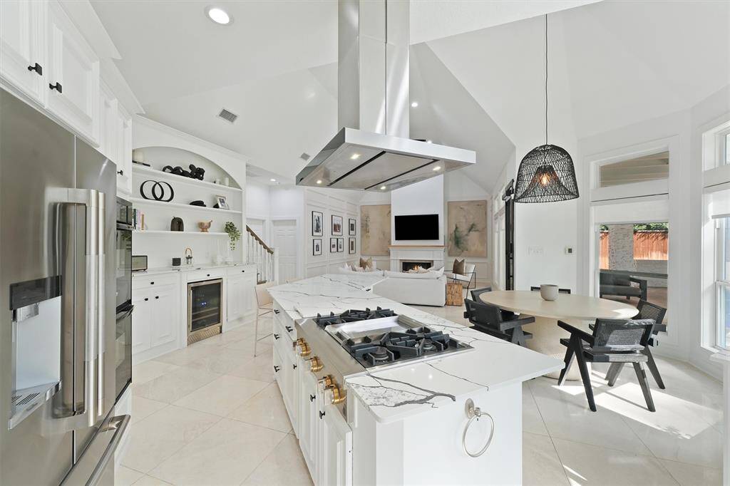 This modern kitchen features sleek white cabinetry, a large island with a gas cooktop, and stainless steel appliances. It opens to a bright living area with high ceilings, a fireplace, and ample natural light from large windows. Perfect for entertaining and family gatherings.