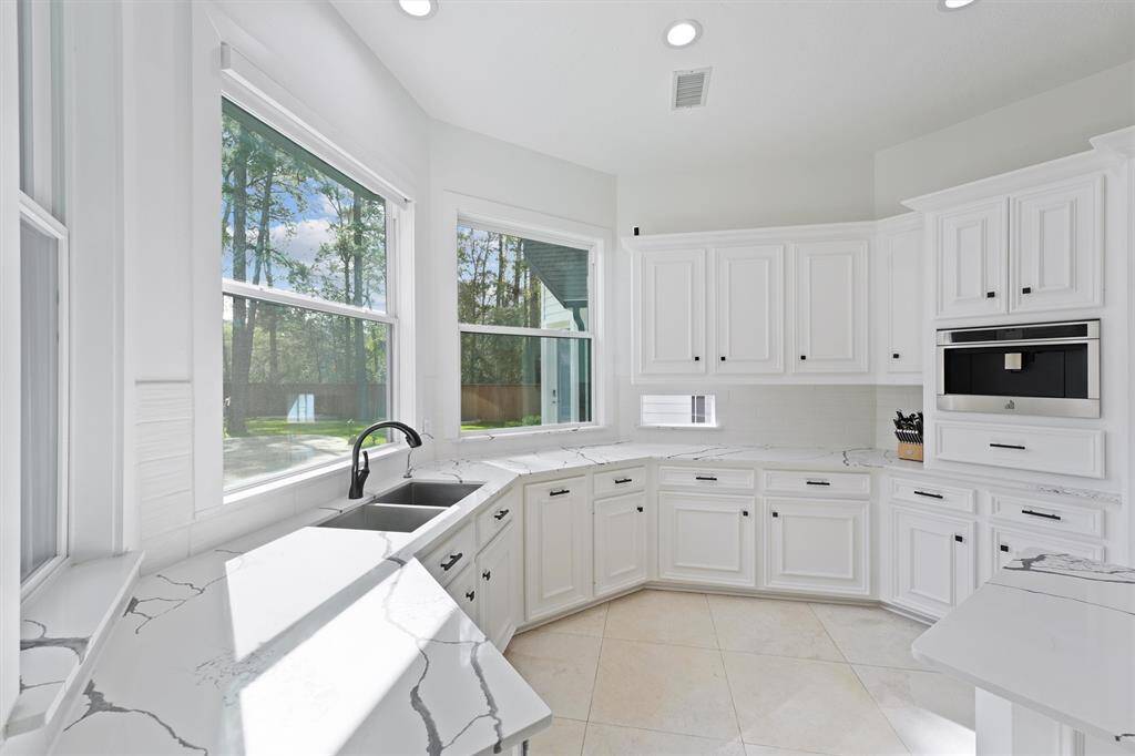 Bright, modern kitchen with large windows offering plenty of natural light. Features white cabinetry, sleek countertops, and built-in appliances. The view overlooks a serene outdoor area, creating a welcoming and airy atmosphere.