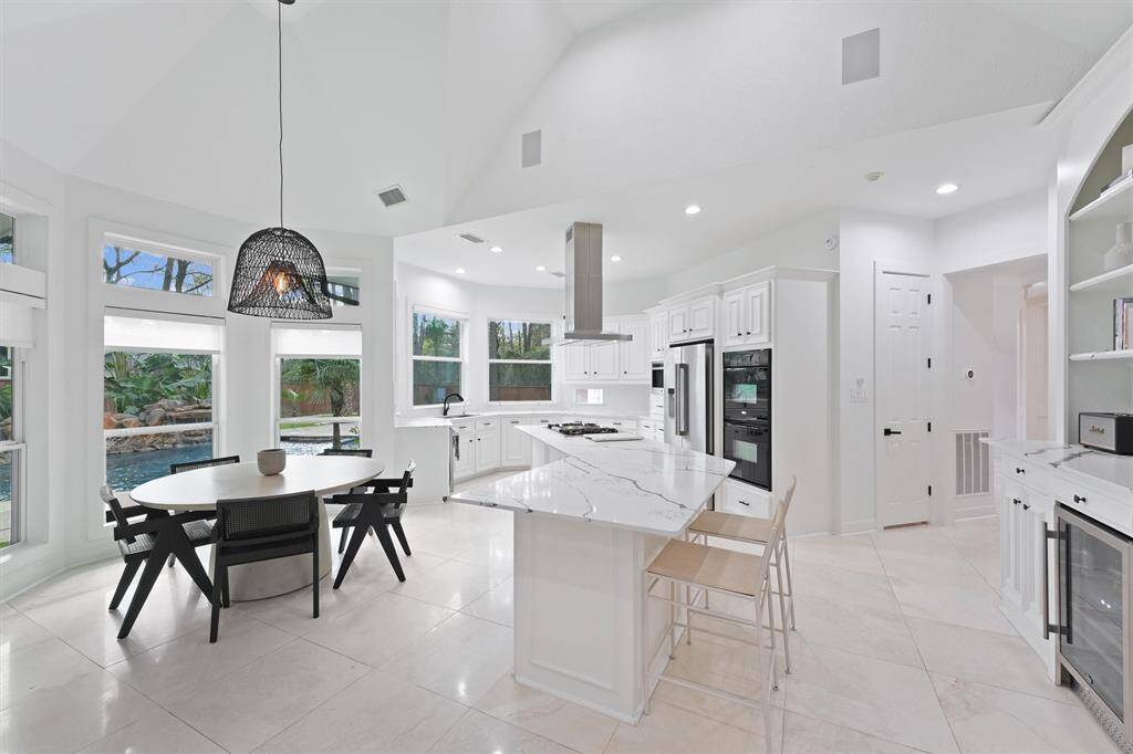 This bright, modern kitchen features a spacious island with marble countertops, sleek white cabinetry, and built-in appliances. Large windows provide ample natural light and views of a lush backyard with a pool. A cozy dining area with a stylish pendant light completes the space.