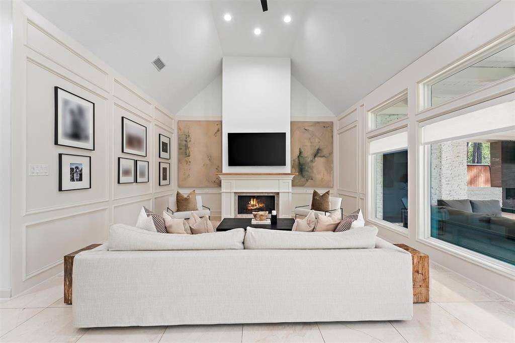This spacious living room features high vaulted ceilings, an elegant fireplace, large windows for ample natural light, and stylish wall paneling adorned with modern artwork. A comfortable, neutral-toned sofa set creates an inviting atmosphere.