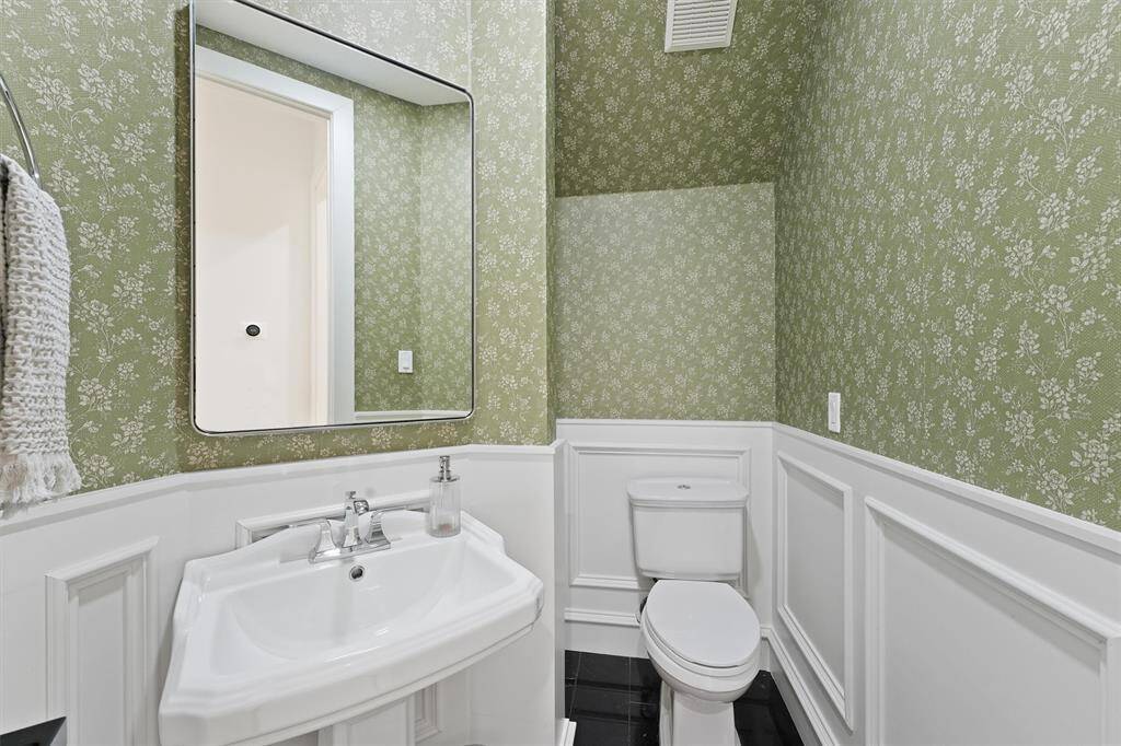 This bathroom features elegant green floral wallpaper, a white wainscot panel, and a sleek pedestal sink with a large mirror, creating a charming and classic look.