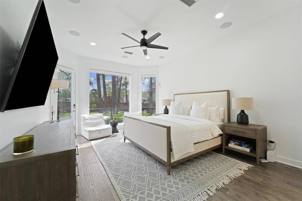 This spacious bedroom features large windows offering abundant natural light, a modern ceiling fan, and elegant decor. It includes a cozy seating area, stylish furniture, and a view of the backyard, creating a serene and inviting atmosphere.