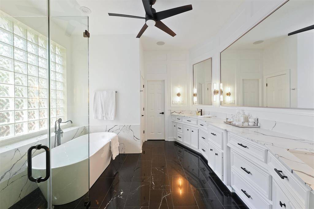This bathroom features a sleek, modern design with a freestanding tub next to a large glass-block window. It has dual vanities with marble countertops, elegant black tile flooring, and a spacious glass-enclosed shower. The room is accented by a stylish ceiling fan and ample natural light.
