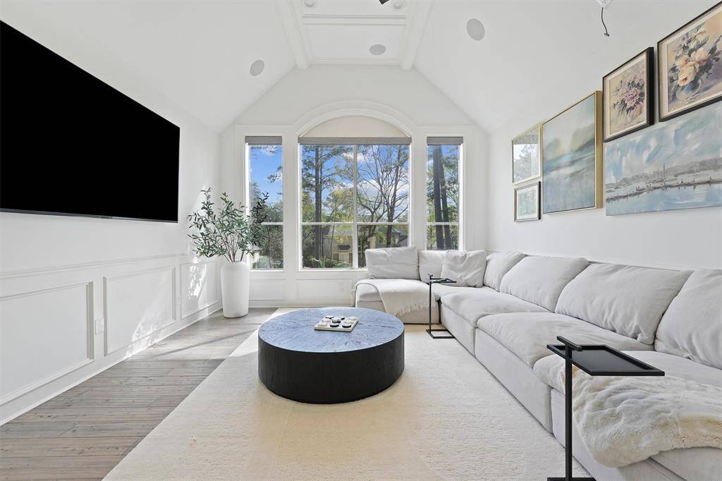 Bright, modern game room with vaulted ceiling and large windows offering natural light and scenic views. Features a spacious, plush sectional sofa, a large wall-mounted TV, and tasteful artwork. Ideal for relaxation and entertainment.
