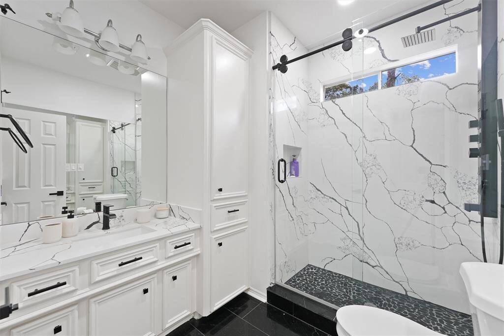 This modern bathroom features a sleek design with a large walk-in shower showcasing elegant marble-patterned tiles. It includes a spacious vanity with ample storage, a wide mirror, and contemporary fixtures, creating a bright and stylish space.
