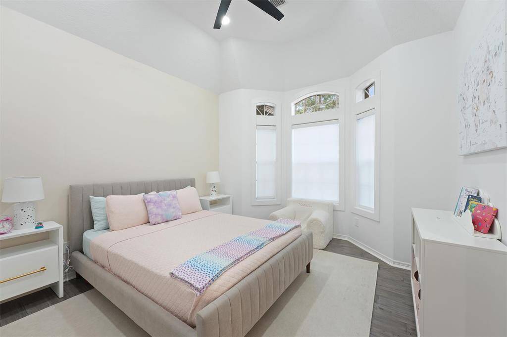 This bright bedroom features a cozy bed with pastel accents, large windows with ample natural light, and modern decor. It includes nightstands, a dresser, and a soft rug on a dark wood floor.