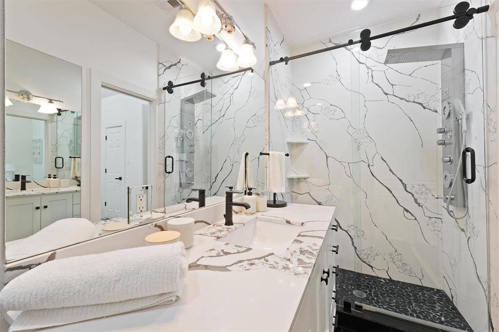 This modern bathroom features a sleek double vanity with marble countertops and elegant black fixtures. The spacious shower boasts a glass door and luxurious rainfall showerhead, complemented by stylish wall tiles and ample lighting.