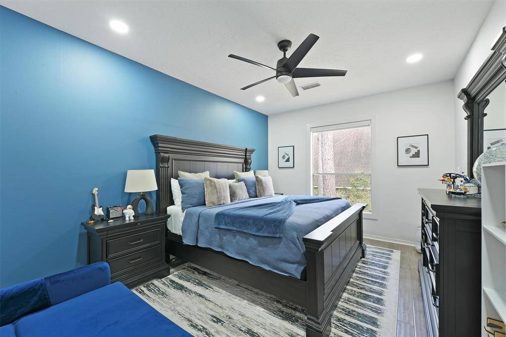 This bedroom features a modern design with a bold blue accent wall and sleek dark wood furniture. It has a large bed with plush bedding, a ceiling fan, and ample natural light from a window. The room is stylishly decorated with a combination of contemporary artwork and accessories.