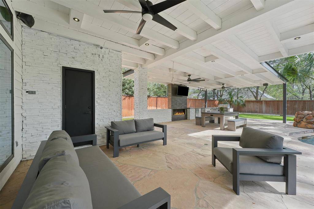 This photo showcases a spacious, covered outdoor patio with modern seating, a ceiling fan, and a built-in fireplace. It features a stone floor, TV, and an outdoor kitchen area, all overlooking a fenced backyard with a pool, creating a perfect space for entertaining and relaxation.
