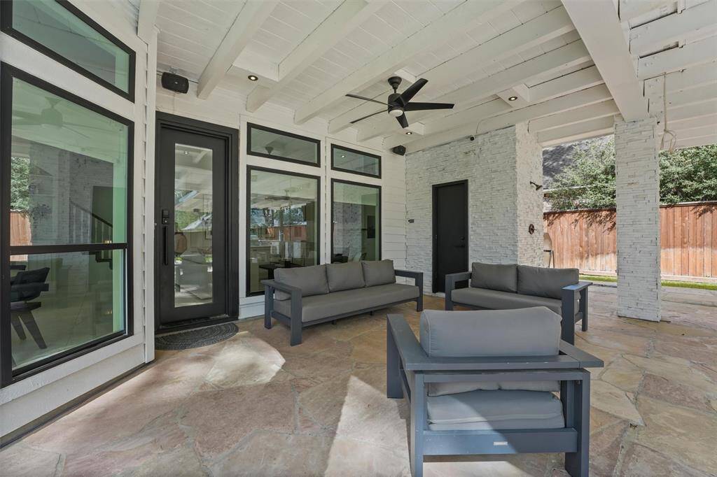 This outdoor patio offers a spacious, covered seating area with modern furniture, a ceiling fan, and stylish stone flooring. Surrounded by large windows and a wooden fence, it provides a comfortable and private space for relaxation or entertaining.