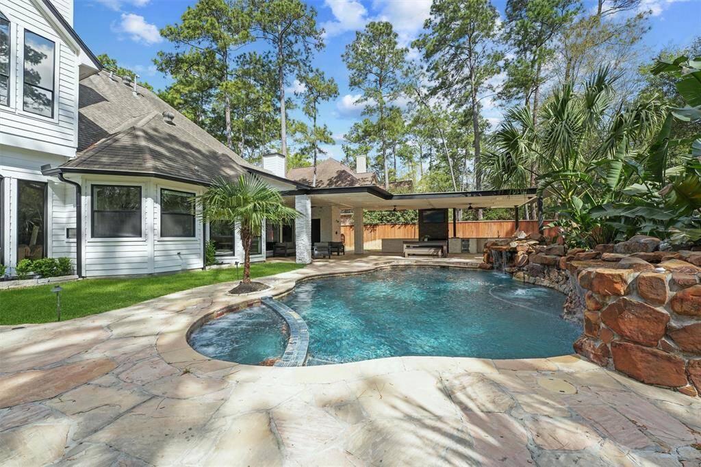 This photo showcases a beautiful backyard with a swimming pool, surrounded by a natural stone patio and lush greenery. There's a covered outdoor living area, perfect for entertaining or relaxing. The home exterior features white siding, large windows, and is nestled among tall trees, offering a serene and private setting.