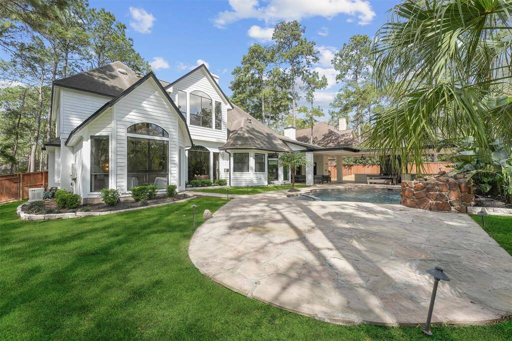 This photo showcases a spacious, modern home with a striking white exterior and large windows, set against a backdrop of tall trees. The backyard features a well-maintained lawn, a stone patio, and a pool, offering a serene and private outdoor space perfect for relaxation and entertaining.