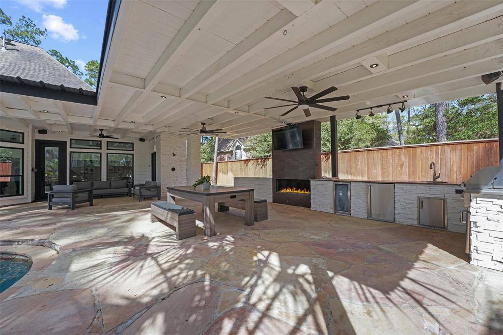 This outdoor space features a covered patio with a ceiling fan, built-in fireplace, and a modern outdoor kitchen area. It includes a spacious dining table, seating, and overlooks a pool, perfect for entertaining. The area is bright and airy, with a wood-paneled fence providing privacy.