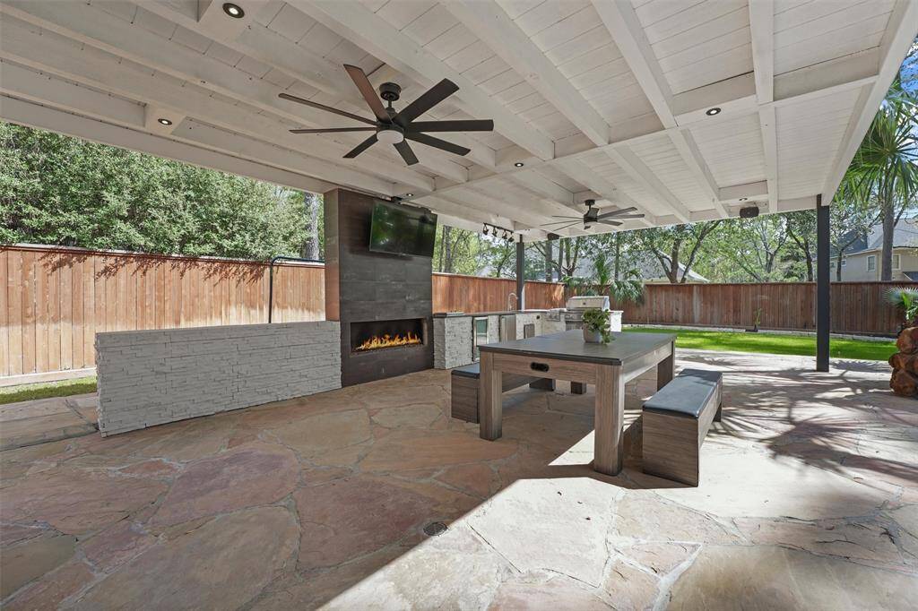 This outdoor space features a covered patio with a modern fireplace, ceiling fans, and a mounted TV. The area includes a large dining table with benches and a built-in grill, all set on a stone floor, overlooking a fenced yard with lush greenery. Perfect for entertaining and relaxing.