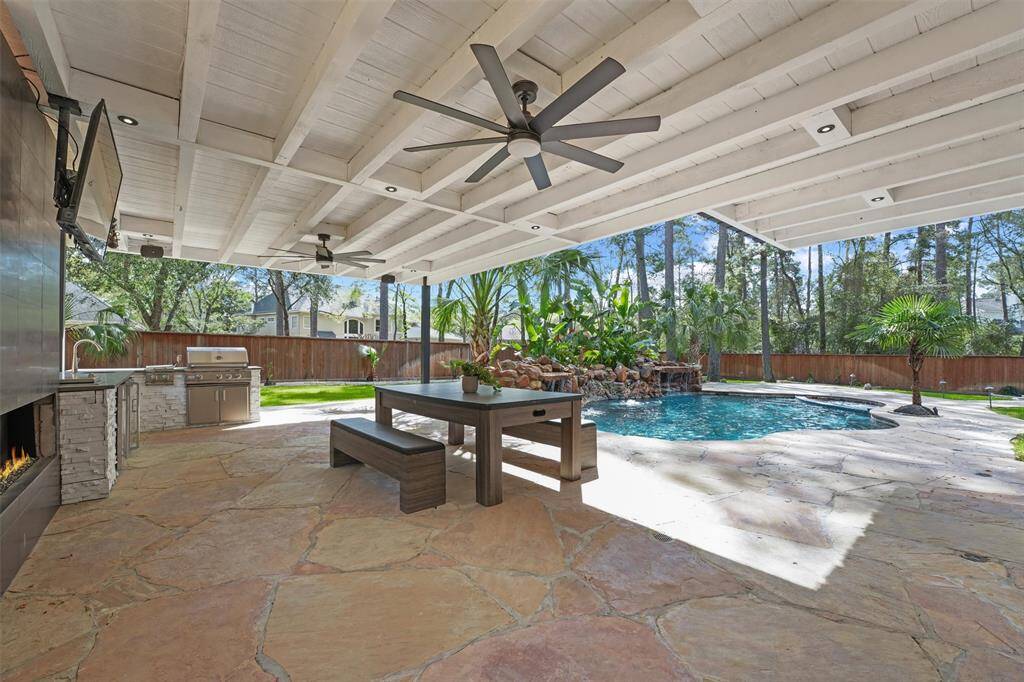 This outdoor space features a spacious covered patio with ceiling fans, a stone fireplace, and an outdoor kitchen with a grill. It overlooks a landscaped backyard with a pool and waterfall, providing a serene and private retreat.