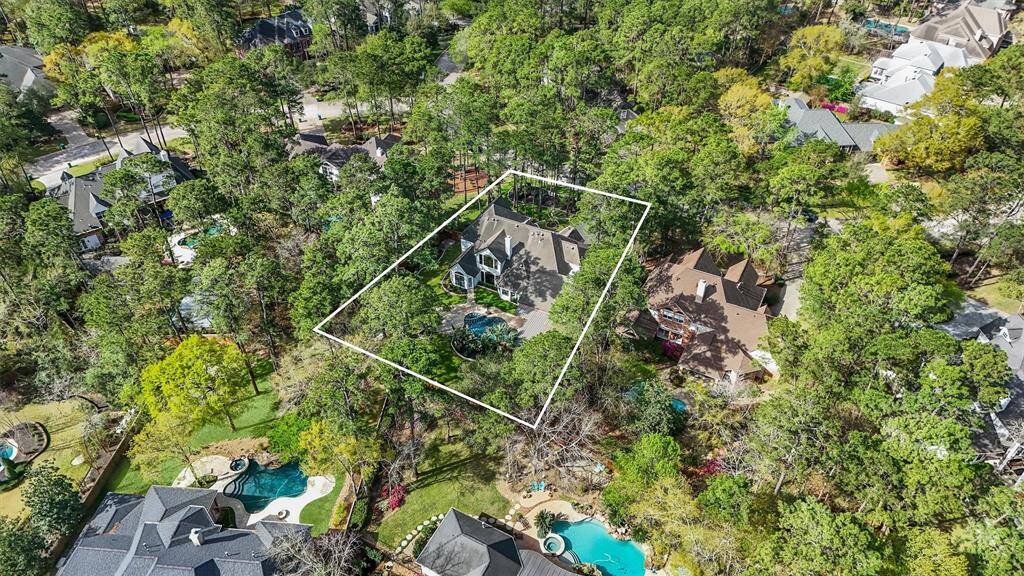 This aerial photo showcases a spacious property outlined in white, nestled in a lush, wooded neighborhood. The home features a large yard with mature trees, offering privacy and natural beauty. The surrounding area includes other well-maintained homes, each with its own unique landscaping and amenities like pools.