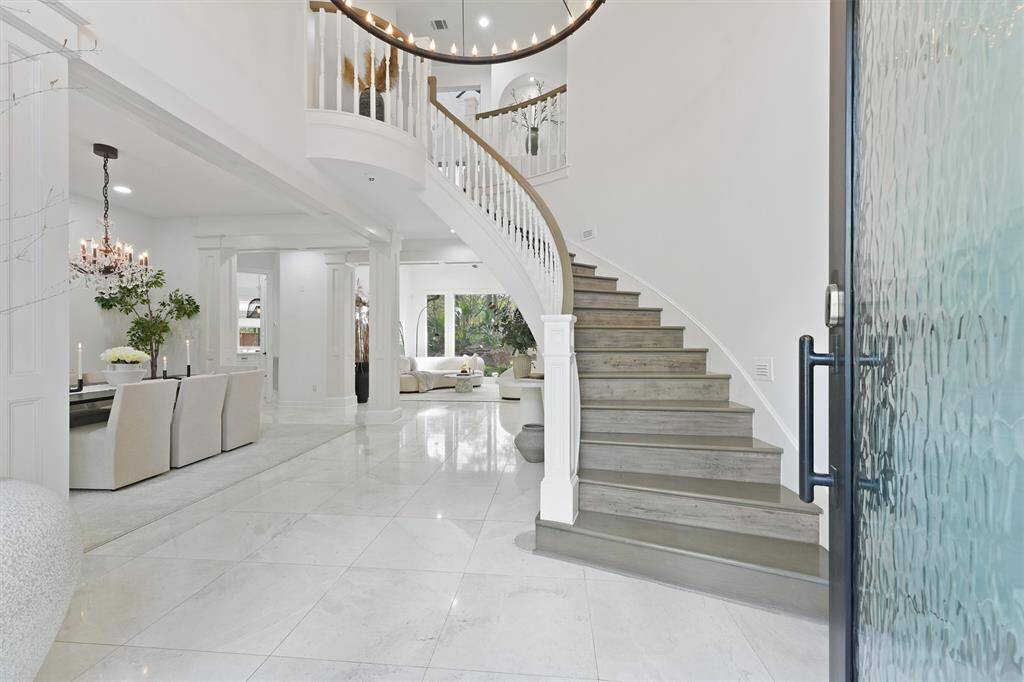 This elegant entryway features a grand spiral staircase, polished travertine floors, and an open layout leading to a formal dining area with a chandelier and a spacious living room. Bright and inviting, perfect for sophisticated living.
