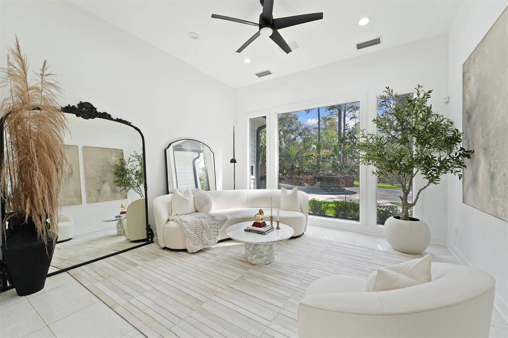 This elegant living room features large windows with a view of lush greenery, sleek white furnishings, and modern decor. It includes a stylish ceiling fan, a large decorative mirror, and tasteful plant accents, creating a bright and inviting atmosphere.