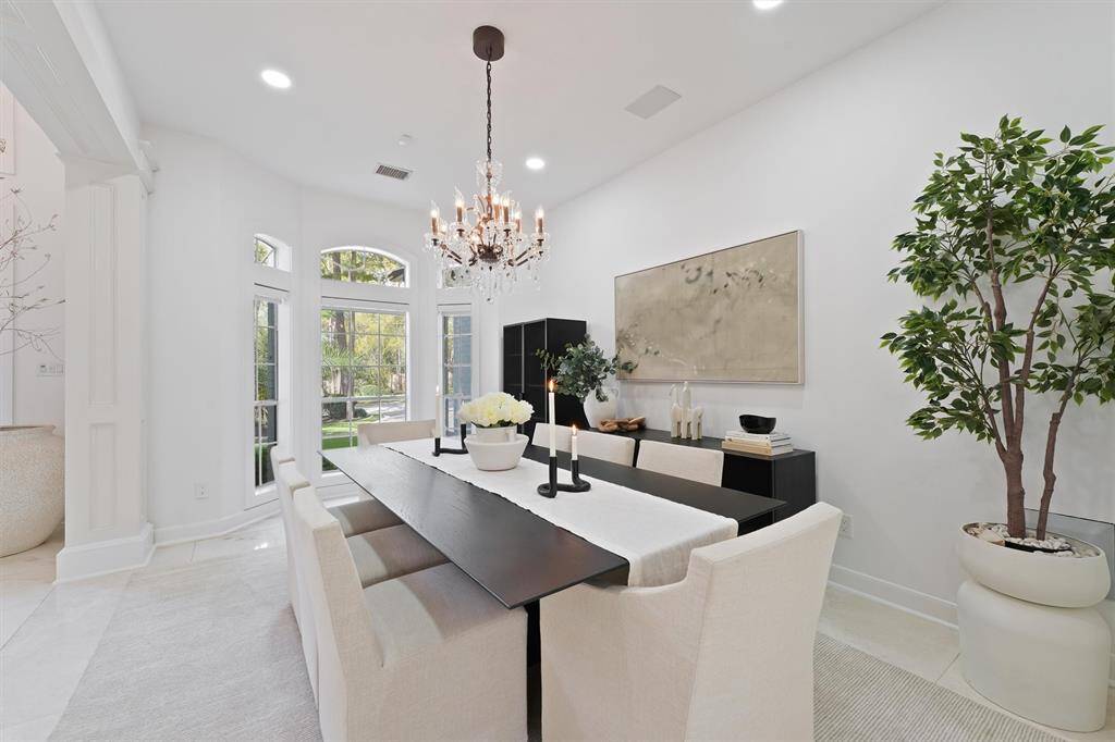 This photo showcases a bright and elegant dining room with a modern chandelier, large windows providing natural light, and a stylish, minimalistic decor featuring a long dining table, comfortable chairs, and tasteful artwork. The room is accented by indoor plants, adding a touch of greenery.