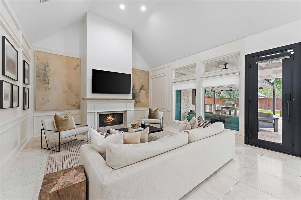 Bright and spacious living room with high ceilings, a modern fireplace, and large windows offering plenty of natural light. The room is elegantly furnished with a neutral color palette, and there's a view of the outdoor patio through glass doors.