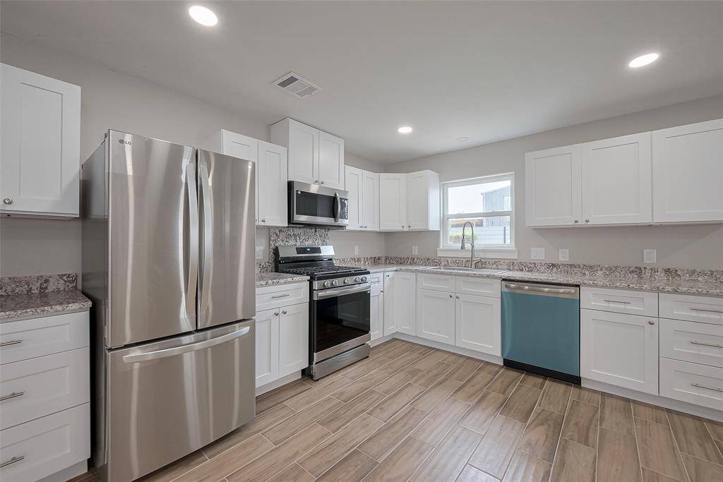 This kitchen features modern stainless steel appliances, white cabinetry, and granite countertops. The wood-look tile flooring adds warmth, and ample natural light brightens the space.