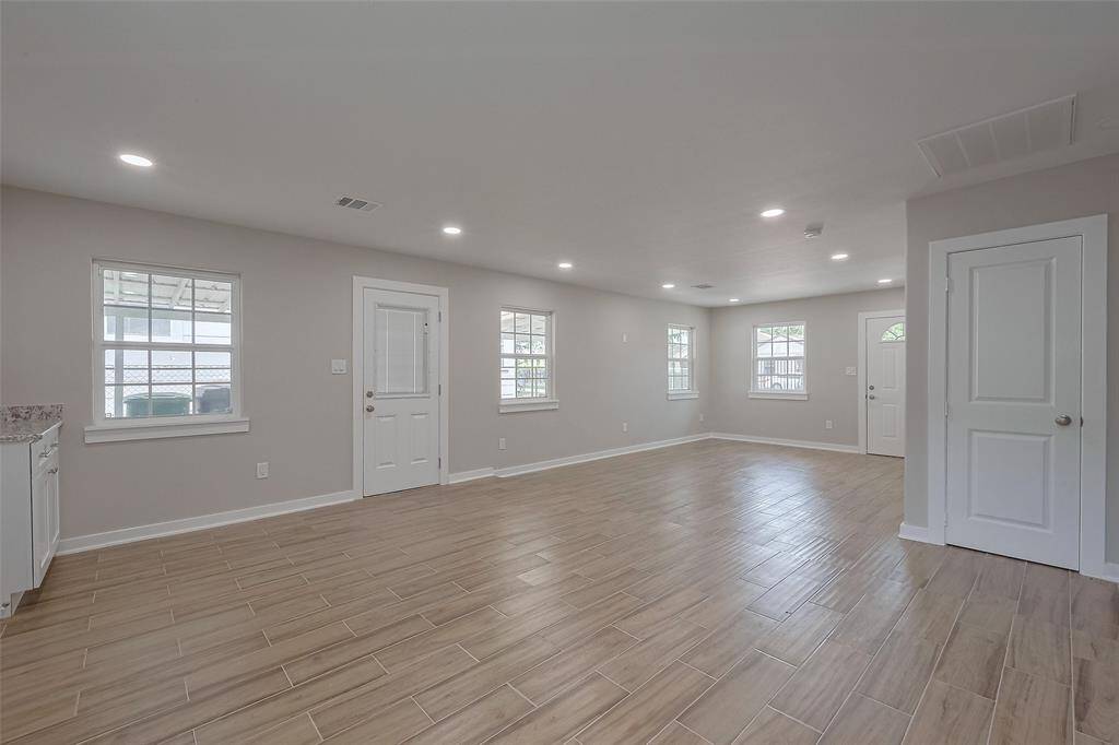 This photo showcases a spacious, open-concept room with wood-like tile flooring, neutral walls, and recessed lighting. Multiple windows provide ample natural light, and there are two doors leading to different areas, enhancing accessibility.