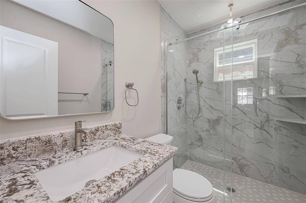 This modern bathroom features a sleek design with a marble-patterned walk-in shower, glass doors, and a rainfall showerhead. The vanity has a granite countertop with a contemporary faucet and a large, stylish mirror.