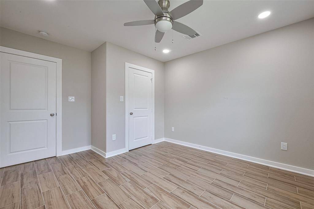 This is a modern, empty room with neutral walls and wood-look tile flooring. It features a ceiling fan with lights and recessed lighting, providing a bright and airy feel. Two white doors add to the clean, minimalistic design.