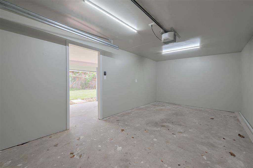 This photo shows a clean, empty garage with light gray walls and a concrete floor. The space is well-lit with fluorescent lights and features a garage door opener. There's a doorway leading to a grassy outdoor area.