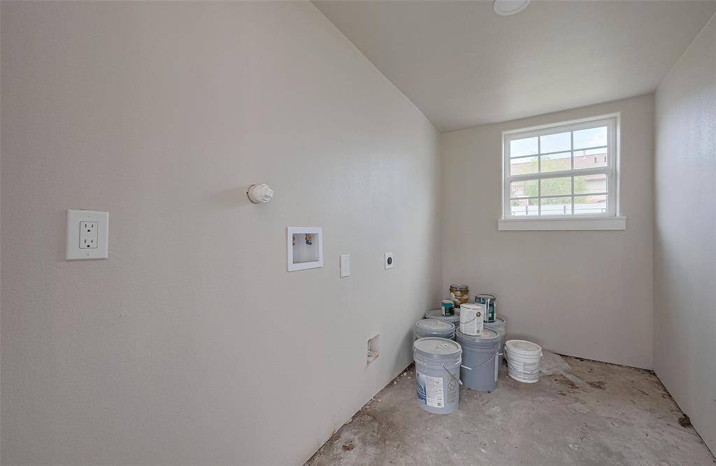 This photo shows a utility room with a window, hookups for a washer and dryer, and a concrete floor. Paint buckets are stored in the corner, suggesting ongoing renovations.