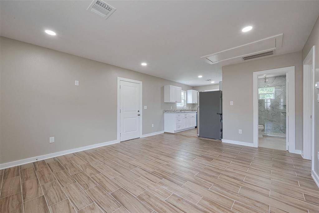 This photo showcases a modern, open-concept living area with wood-look tile flooring and recessed lighting. The space flows into a bright kitchen with white cabinetry and granite countertops, and there's a glimpse of a contemporary bathroom with a tiled shower.