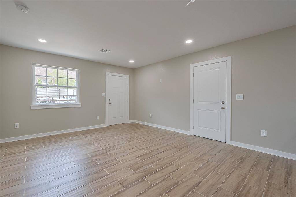 This photo shows a bright, unfurnished room with wood-look tile flooring, neutral-colored walls, recessed lighting, and two doors. A window provides natural light, creating a welcoming atmosphere.