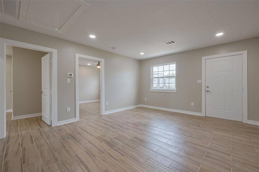 This photo shows a bright, open living area with neutral walls and wood-look tile flooring. It features recessed lighting, a window for natural light, and a door leading outside. The space is versatile and inviting, ideal for personalizing.