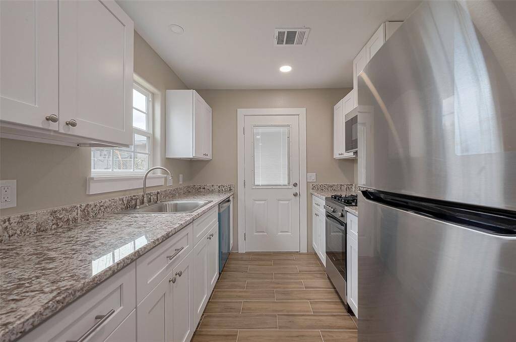 This modern kitchen features sleek stainless steel appliances, granite countertops, and ample white cabinetry. The space has a bright ambiance with natural light from the window and a door leading outside. Wood-look tile flooring adds warmth and elegance.