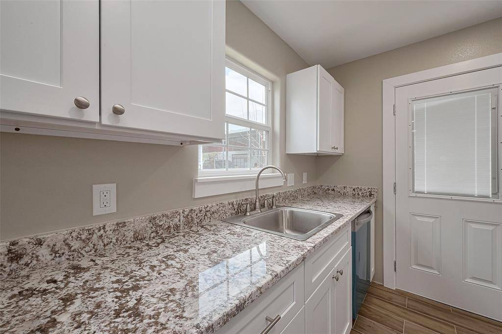 This bright kitchen features granite countertops, a stainless steel sink, and white cabinetry. A large window and a door with a built-in blind provide ample natural light.