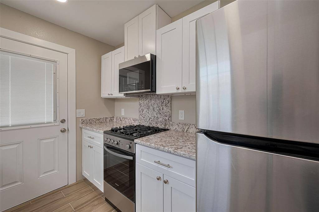 This modern kitchen features sleek stainless steel appliances, including a refrigerator and gas stove, complemented by white cabinetry and granite countertops. The space is bright and functional, with a stylish tile backsplash and a convenient door leading outside.