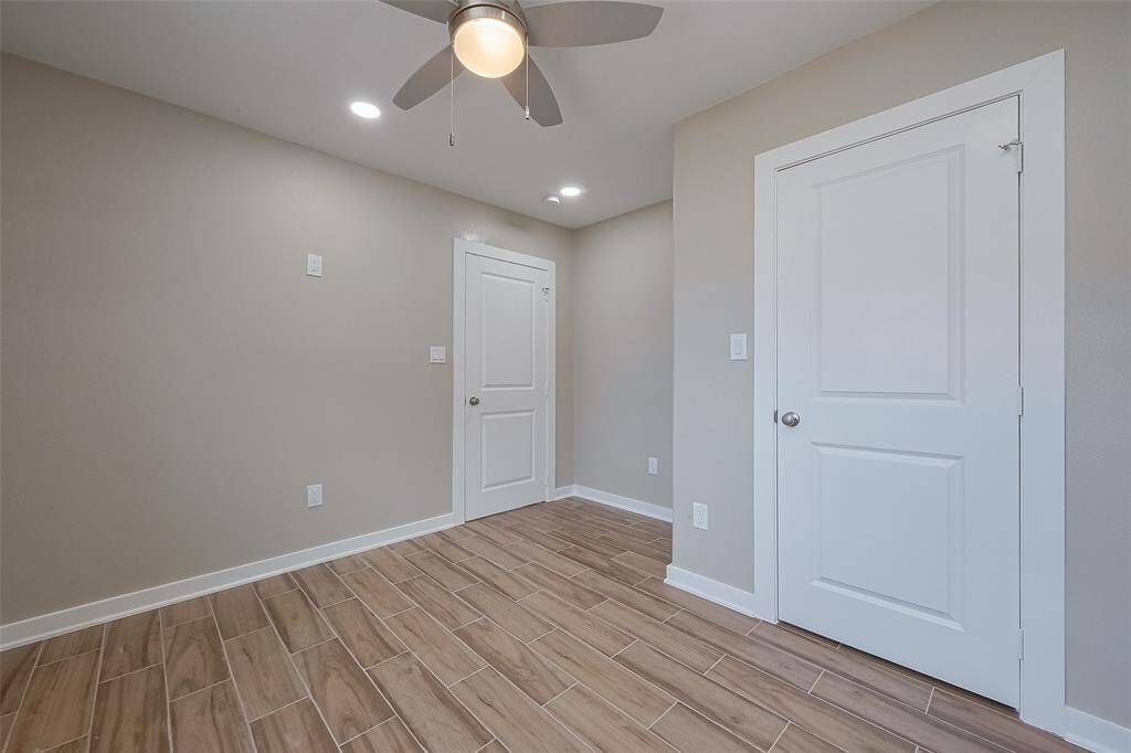 A bright room with light gray walls and wood-look tile flooring. It has a ceiling fan with lights and two white doors, offering a clean and modern feel.