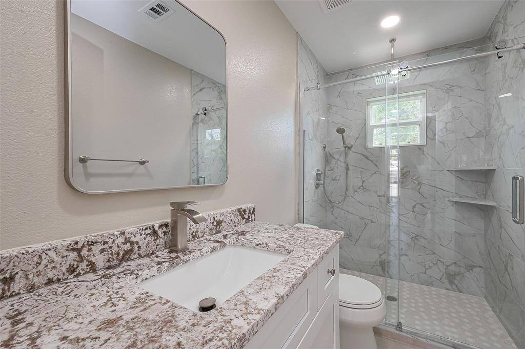 This bathroom features a modern design with a large mirror, sleek granite countertop, and a spacious glass-enclosed shower with marble-patterned tiles.