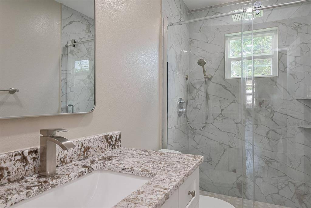 All New bathroom features a modern design with a granite countertop, sleek faucet, and a spacious glass-enclosed shower with marble-patterned tiles. Natural light streams in through a window, enhancing the elegant atmosphere.
