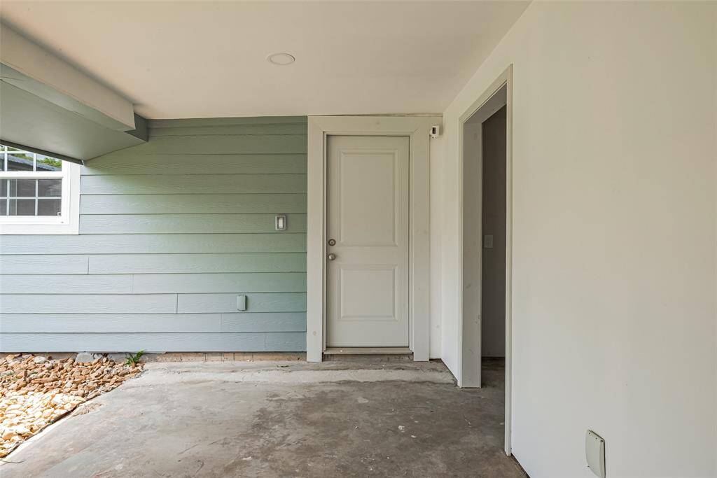 Covered entryway of the guest house with a light blue exterior wall and a white door. The space has a partially exposed concrete floor and a small section of landscaping with stones.